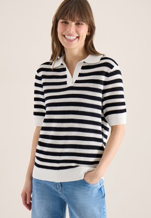 Kobieta uśmiechająca się, ubrana w krótki rękaw biało-czarny pasiasty sweter z kołnierzykiem i jasnoniebieskie dżinsy, ręka w kieszeni, na neutralnym tle.