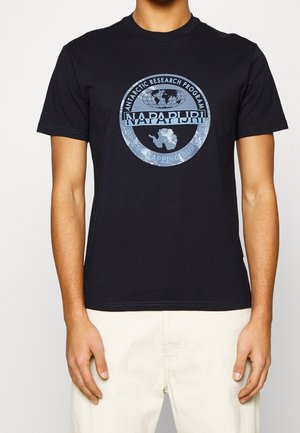Mann trägt schwarzes kurzärmeliges T-Shirt mit blauem rundem Logo des Antarctic Research Program und weiße Hose, steht vor einfarbigem Hintergrund.