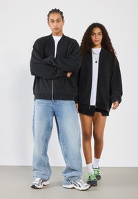 Black zip-up bomber jackets, loose blue jeans, and athletic shoes. The woman wears a black zip-up jacket and shorts, both casual and relaxed.