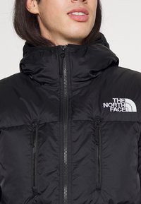 Casaco preto acolchoado com um colarinho alto, fecho zip e dois bolsos com fecho zip. Apresenta o logo "The North Face" no peito.