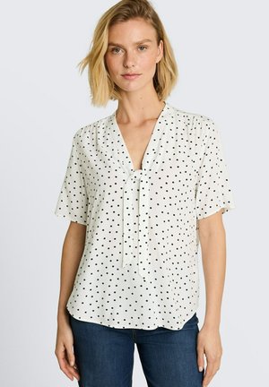 Femme blonde portant un chemisier blanc à manches courtes avec des pois noirs et un col à nouer, associé à un jean bleu foncé, debout devant un fond uni.