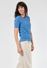 Vaaleansininen neulottu lyhythihainen neulepaita, jossa on teksturoitu timanttikuvio ja ribbauskaulus, yhdistettynä vaaleanbeigeihin housuihin.