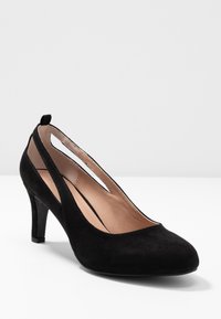 Escarpin pour femme en daim noir à talon bas, bout pointu, côtés ouverts et doublure intérieure beige, posé sur une surface blanche.