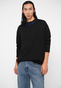 Črn pleten pulover z rebrastimi manšetami in okroglim izrezom, z modrim vezenjem okrog izreza. Nošen z modrimi traper hlačami.