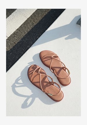 Ein Paar hellbraune flache Riemchensandalen auf einer weißen Oberfläche neben strukturiertem dunklem Asphalt mit einer weißen aufgemalten Linie.