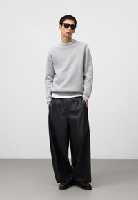 Lichtgrijze gebreide trui, brede zwarte broek en zwarte zonnebril. Losse pasvorm en minimalistisch ontwerp met een effen achtergrond.