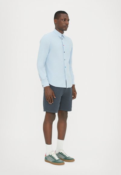 Ljusblå långärmad skjorta med knappframsida, tillsammans med mörka shorts. Har färgglada knappdetaljer och gröna sneakers med vita strumpor.