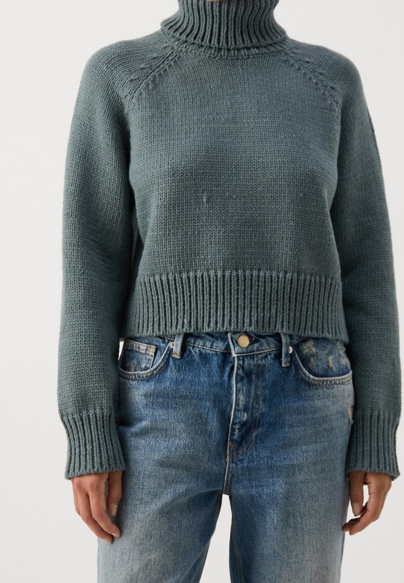 Personne portant un pull à col roulé tricoté couleur sarcelle, rentré dans un jean bleu délavé taille haute, les mains détendues le long du corps.