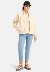 Chaqueta de forro polar en color crema, leggings en azul claro y zapatillas multicolores. Los accesorios incluyen una gorra negra y calcetines beige. Detalles visibles.