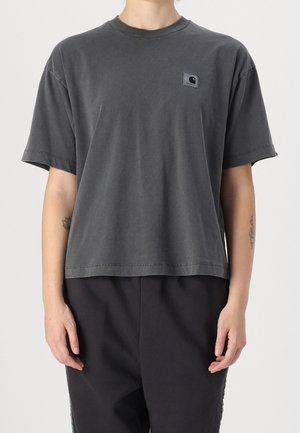 Person trägt ein locker sitzendes dunkelgraues Carhartt-T-Shirt und schwarze Hose und steht vor einem schlichten hellen Hintergrund.