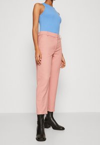 Blaues geripptes Tanktop, kombiniert mit rosa taillierten Hosen und schwarzen Knöchelstiefeln. Das Outfit zeichnet sich durch klare Linien und eine figurbetonte Silhouette aus.