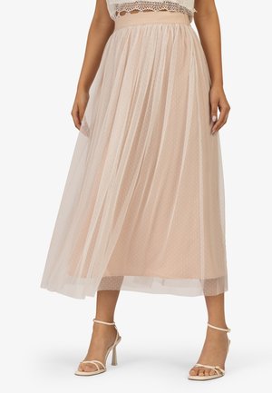 Femme portant une jupe midi beige avec une superposition en tulle transparent à pois et des sandales à talons hauts beiges, vue de la taille aux pieds.