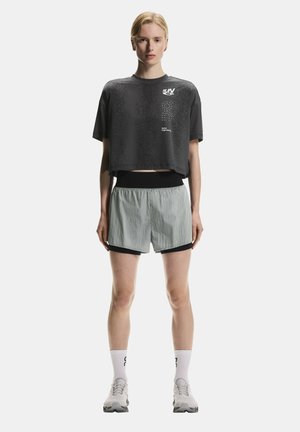 Blonde Frau steht, trägt ein dunkles, kurzes Sportshirt mit Logo, hellgraue, doppellagige Laufshorts, weiße Socken und hellgraue Laufschuhe.