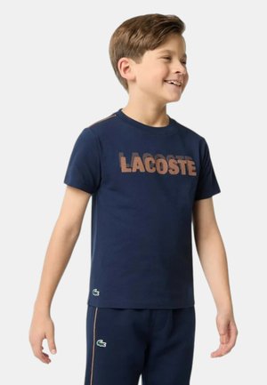 Ragazzo sorridente, indossa una T-shirt Lacoste blu navy e pantaloni abbinati, in piedi davanti a uno sfondo chiaro e uniforme.