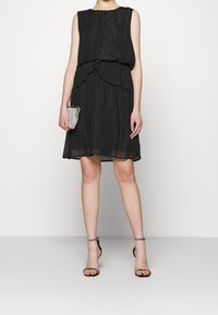 Schwarzes ärmelloses Kleid mit einer strukturierten, schimmernden Oberfläche und einem rüschendetails an der Taille, kombiniert mit einer kleinen, glitzernden Clutch und Riemchenheels.