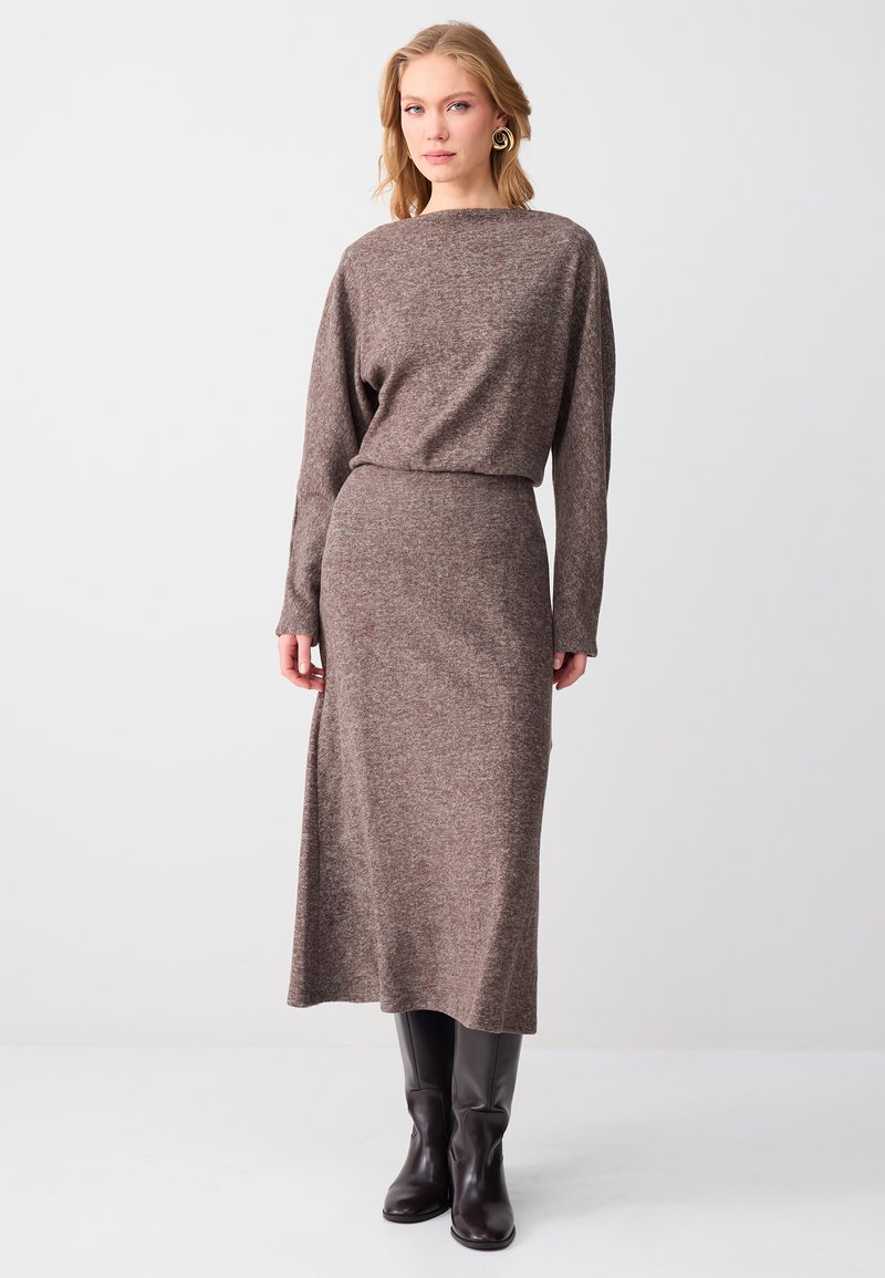 Femme blonde portant une robe en maille marron à manches longues, cintrée à la taille, et des bottes noires montant jusqu'aux genoux, debout devant un fond blanc uni.