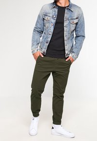 Spijkerjack met lichte wassing, twee borstzakken, zwart T-shirt eronder, olijfgroene joggingsbroek en witte sneakers met zwarte accenten.