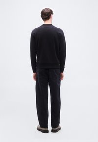 Άνδρας που στέκεται με την πλάτη γυρισμένη, φοράει μαύρο μακρυμάνικο φούτερ, μαύρο τζιν και ανοιχτό καφέ παπούτσια σε λευκό φόντο.