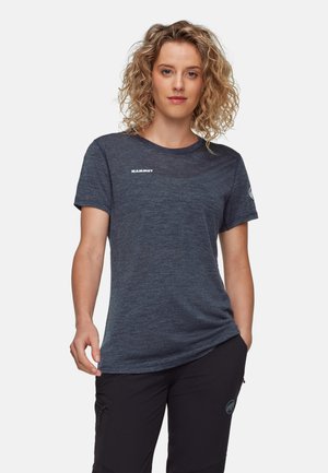 Femme aux cheveux bouclés portant un t-shirt Mammut gris foncé et un pantalon noir, debout avec une main dans la poche sur un fond uni.