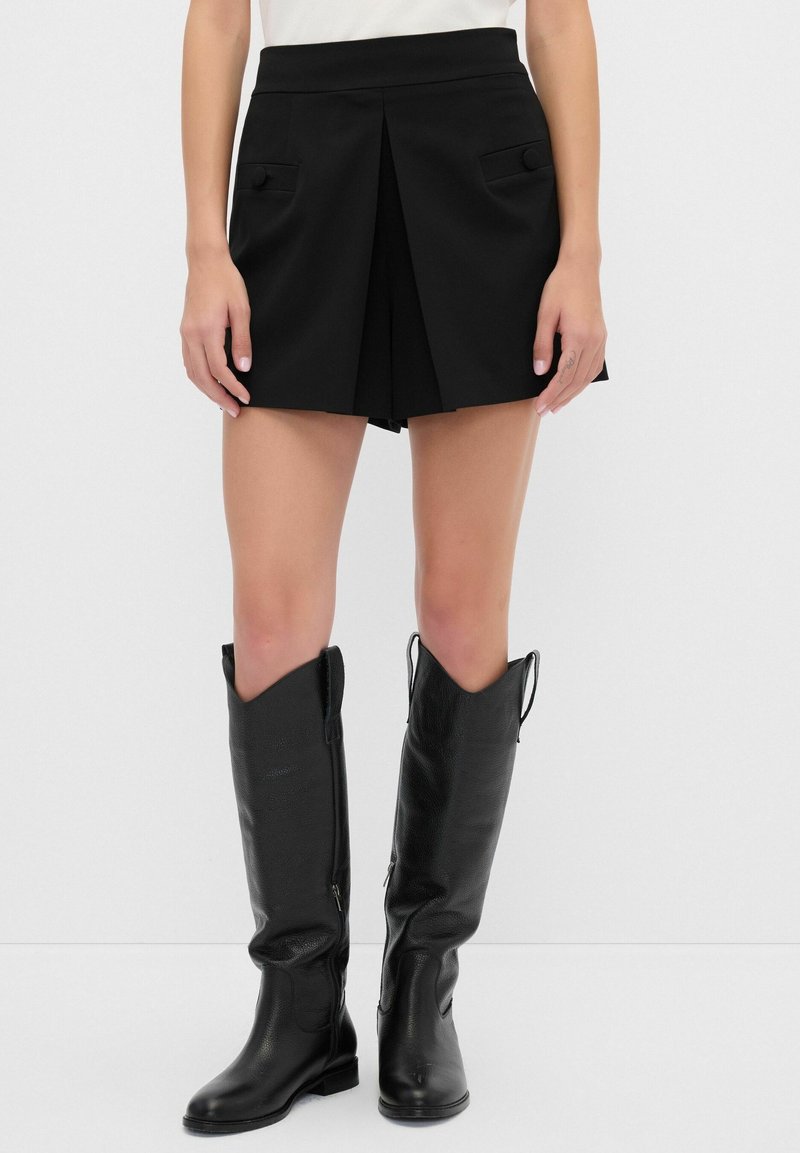 Pantalones cortos negros con un diseño plisado, que presentan bolsillos frontales con botones. El modelo lleva botas altas de cuero negro.