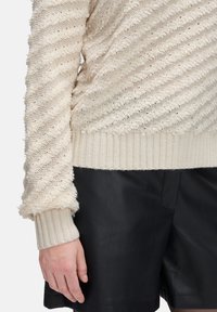 Cremfarbener, strukturiert gestrickter Pullover mit gerippten Bündchen, kombiniert mit schwarzen Ledershorts, die am Oberkörper und Arm einer stehenden Person präsentiert werden.