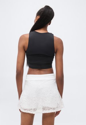 Femeie purtând un top negru fără mâneci și o fustă albă din dantelă, stând cu spatele spre un fundal alb simplu.