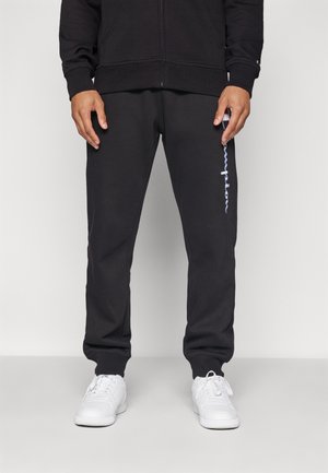 Person iført sorte sweatpants med hvidt lodret logo på venstre lår og hvide sneakers, stående mod grå baggrund.