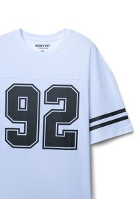 Luźna koszulka z krótkim rękawem w jasnoniebieskim kolorze z bawełny, z czarnym nadrukiem 92 na przodzie, z czarno-białymi paskami na rękawach.