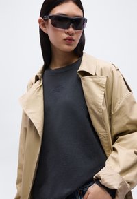 Jeune femme portant un trench-coat beige oversize, un sweat-shirt foncé, un jean et de grandes lunettes de soleil sport noires, se tenant devant un fond uni.
