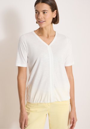 Femme aux cheveux courts et bruns portant un haut blanc à manches courtes avec un col en V et un pantalon jaune clair, regardant vers sa gauche sur un fond neutre.