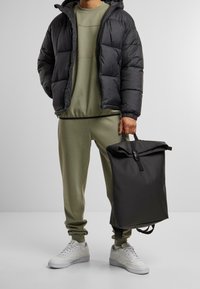 Doudoune noire, sweat-shirt et pantalon vert olive, baskets blanches. Sac à dos noir à rabat roulé avec une sangle tenue dans la main droite.