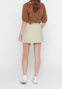 Mini gonna khaki con taglio dritto, abbinata a un top marrone e sneakers alte bianche, dotata di maniche a sbuffo corte e texture liscia.