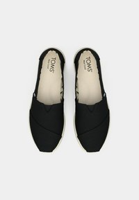 Svarte slip-on sko laget av kanvas med teksturert overflater, med en lyseventet såle og et mønstret innersåle. Merkevaren er synlig.