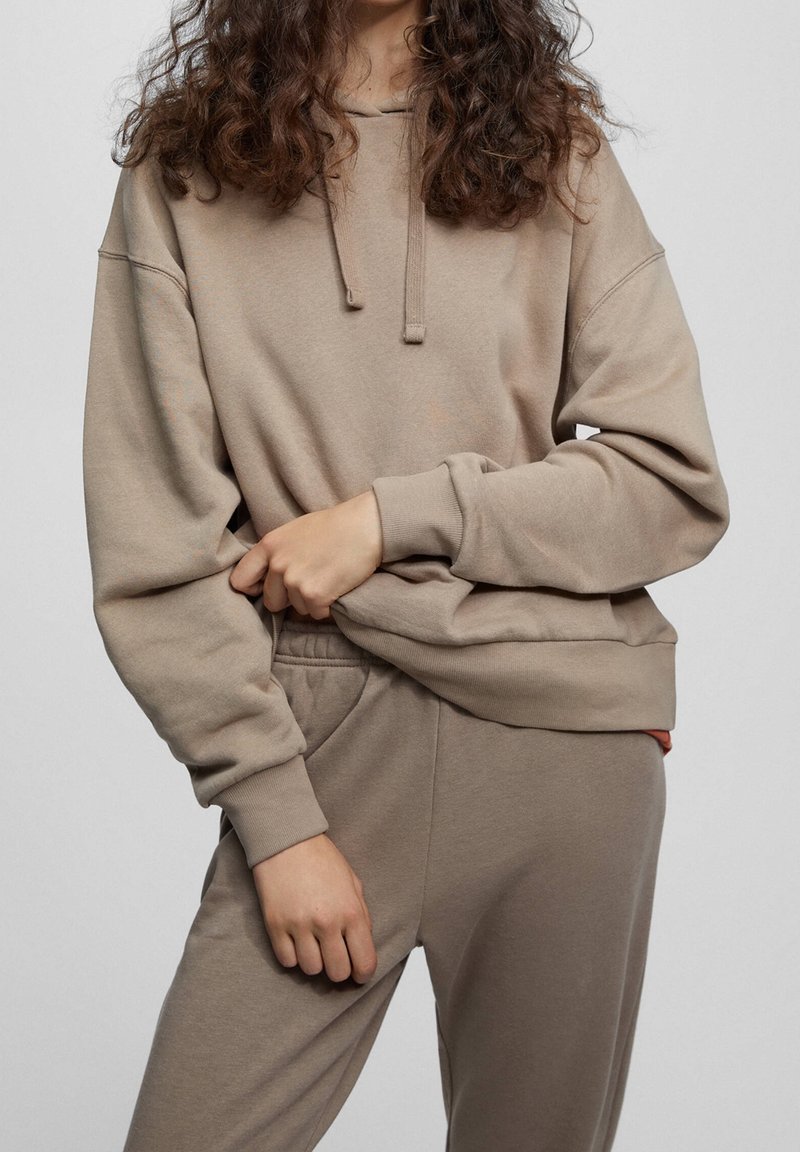 Personne portant un sweat à capuche beige et un jogging assorti, debout, une main soulevant légèrement la capuche, l'autre reposant sur la cuisse.