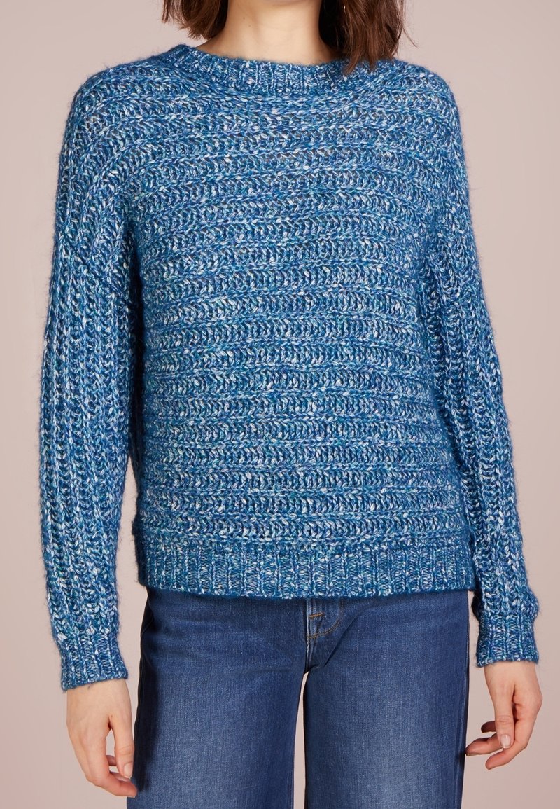 Femme portant un pull en maille texturée bleu et un jean bleu, debout devant un arrière-plan beige uni.