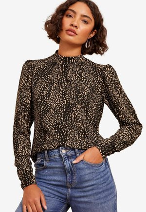 Sort bluse med høj hals og lange ærmer, prydet med et beige spraglet mønster og elastiske ærmer. Bæres med blå denimjeans.