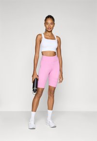 Un crop top alb sportiv și pantaloni scurți roz cu talie înaltă, confecționați dintr-un material neted. O sticlă de apă neagră este ținută în mâna dreaptă.