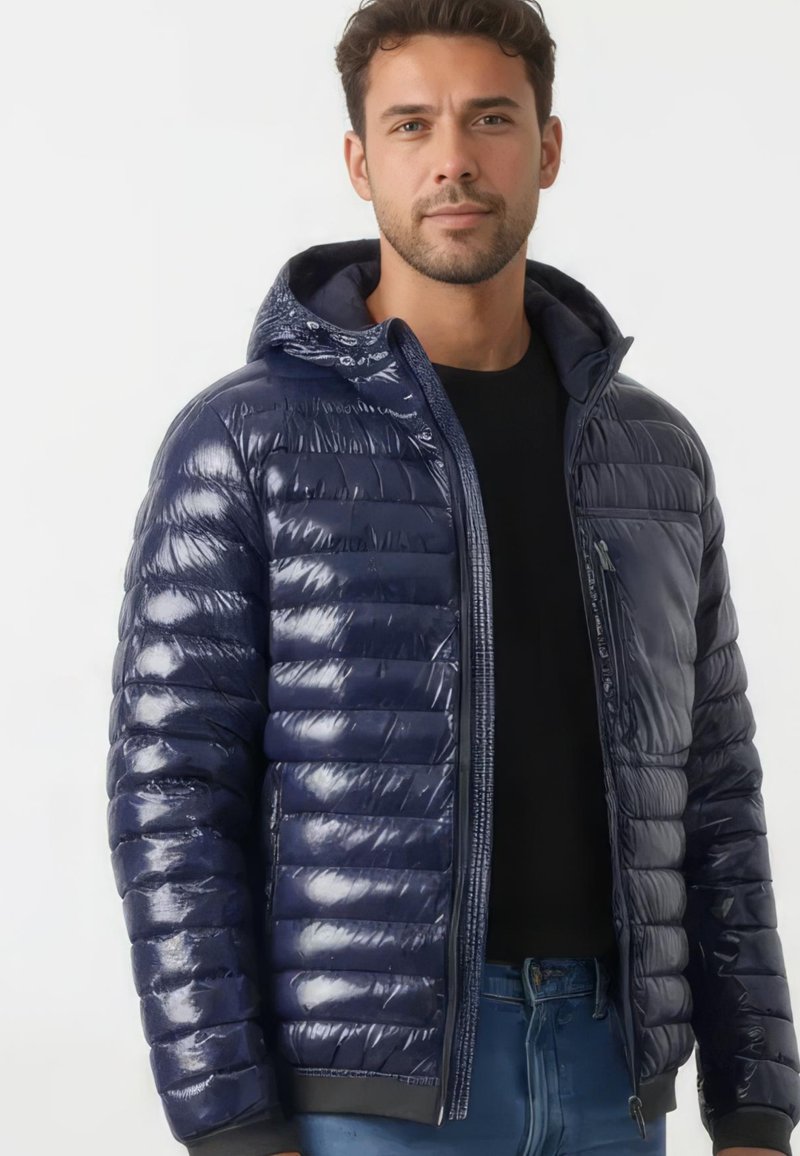 Mann trägt eine glänzende marineblaue Steppjacke mit Kapuze, schwarzes Hemd und blaue Jeans, steht vor einem einfarbigen hellen Hintergrund.