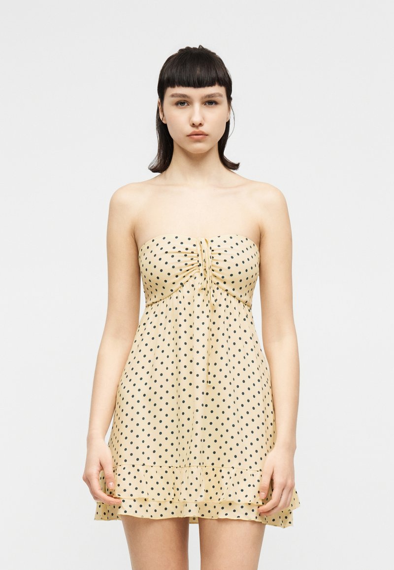 Νέα γυναίκα με κοντά μαύρα μαλλιά, ντυμένη με ένα αμάνικο, ανοιχτό κίτρινο φόρεμα με μαύρες πουά, που στέκεται μπροστά σε ένα απλό λευκό φόντο.