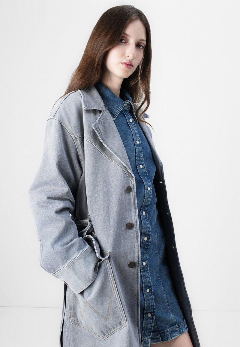 Denim-Mantel in Hellgrau mit strukturiertem Design, ausgestattet mit großen Taschen, Knopfverschlüssen und einem darunter getragenen, mehrlagigen blauen Denim-Hemd.