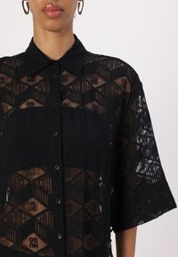 Schwarze durchsichtige Hemdbluse mit Kragen und kurzen Ärmeln, die ein geometrisches Muster des Markenlogos aufweist.