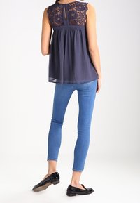 Top azul marino con espalda de encaje, ajuste holgado, diseño en capas; combinado con vaqueros ajustados azul claro y zapatos planos negros. Fondo neutro.