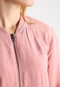 Jasnoróżowa kurtka bombowiec z zamkiem błyskawicznym z przodu, prążkowanym kołnierzem oraz subtelnym marszczeniem na ramionach. Gładka faktura materiału.