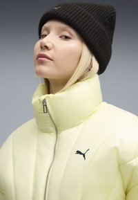Gul puffjacka med hög krage och svart dragkedja. Har en svart Puma-logotyp på vänster sida. Bärs med en svart stickad mössa.