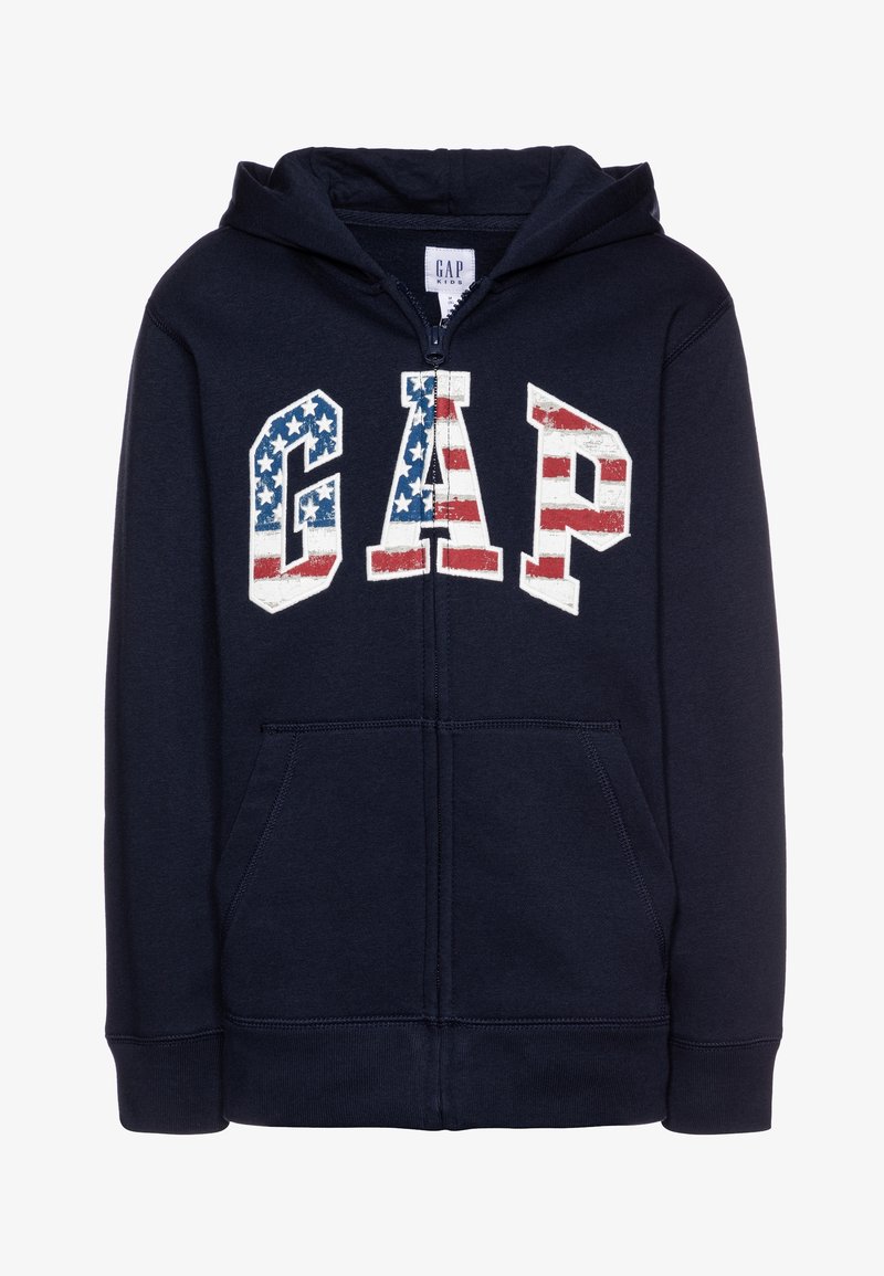 Sweat à capuche zippé bleu marine en tissu doux, arborant un grand logo "GAP" avec un motif à rayures rouges, blanches et bleues. Deux poches avant.