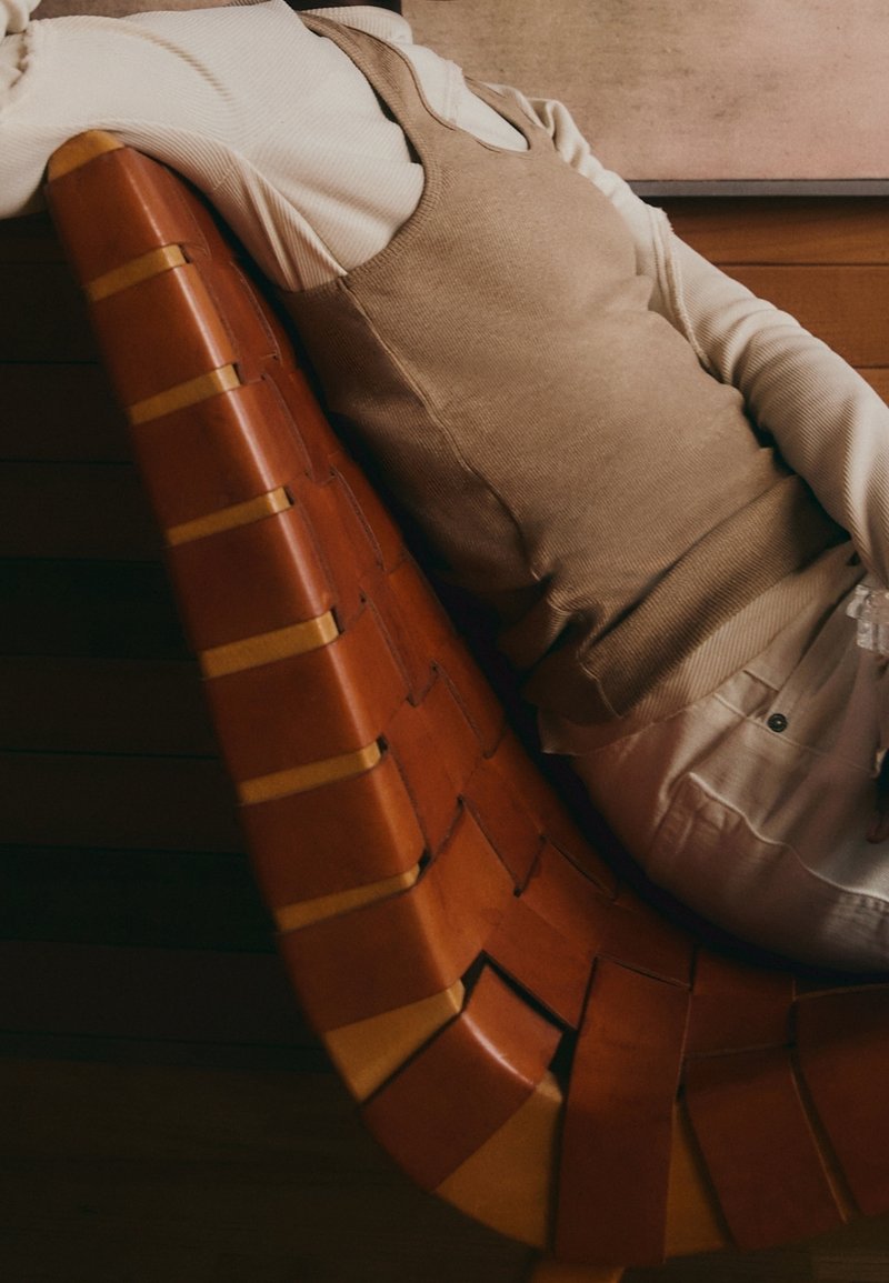 Personne portant un débardeur beige et une chemise blanche à manches longues, allongée sur une chaise en cuir marron tressé avec un sol en bois et un mur en arrière-plan.