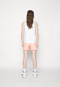 Bílý bezrukávový top, broskvové šortky, bílé vysoké tenisky s červenými akcenty, model viděný zezadu. Jednoduché pozadí.