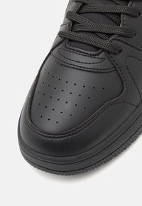 Gros plan de la partie avant d'une basket en cuir noir avec des perforations et des lacets noirs sur fond blanc.