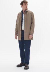 Tan waterproof coat with a stand-up collar, button front, paired with a blue button-up shirt and dark jeans. Model wears brown shoes.