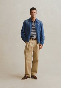 Άνδρας με μπλε μπουφάν, πουκάμισο με μπλε και λευκές ρίγες, μπεζ παντελόνι και καφέ παπούτσια, στέκεται μπροστά σε απλό φόντο.